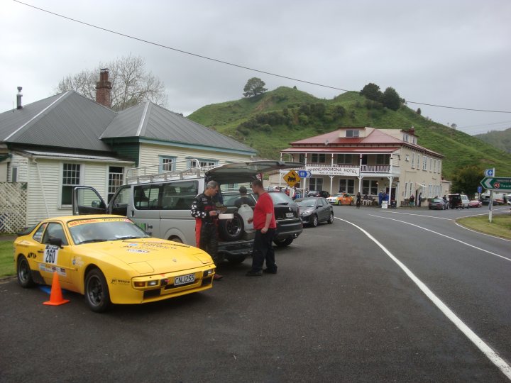 Targa 2013 - Page 1 - New Zealand - PistonHeads - The image depicts a group of people standing in a parking lot, seemingly inspecting or admiring a pair of vintage sports cars. The cars are yellow and appear to be a mix of a coupe and a convertible model. They are parked side-by-side and take up a significant portion of the foreground. In the background, there are buildings and cars, indicating that the parking lot might be in an urban area. The scene is featureless except for the two cars, and the general atmosphere seems to be one of interest or authority regarding the cars.