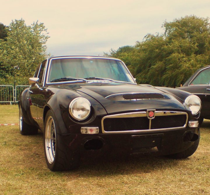 Pistonheads - The image showcases a well-maintained black MG sports car parked on a grass-covered field. The vehicle features a distinctive, pointed front end, with headlights that flare outward, and a large radiator grille. It is equipped with a black soft top, just in front of which the words "MG" are proudly displayed. In the background, there is a line of cars parked at a slight angle, and part of a tree and a green fence is visible. The sky overhead is overcast with no signs of precipitation. The overall scene suggests a gathering of classic car enthusiasts.