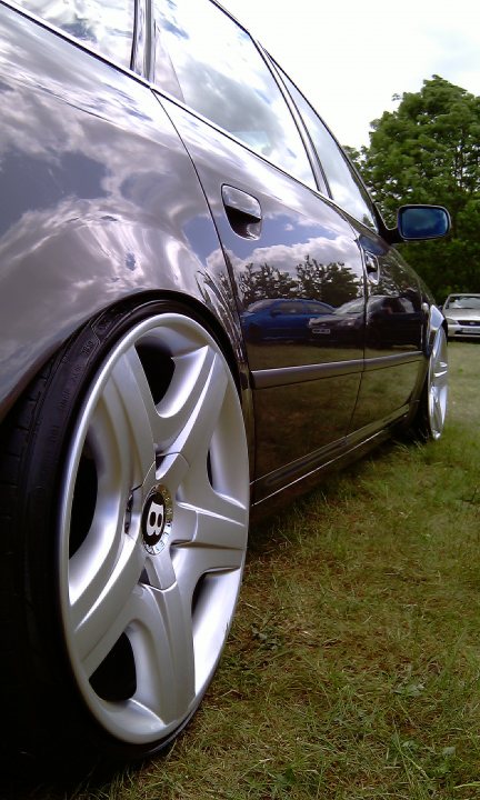 Cars On The Green - Photo Thread - Page 1 - East Anglia - PistonHeads - The image showcases the front left wheel of a black car. The chrome hubcap and silver rims catch the light, contrasting with the car's shiny surface. The car is parked on a grassy area, with its reflection visibly distorted on the hood due to sunlight and a wet surface. In the background, another car can be seen parked in the distance. The image creates a dynamic interplay between the stillness of the parked vehicle and the activity of the presupposedly wet surface reflecting the surroundings.