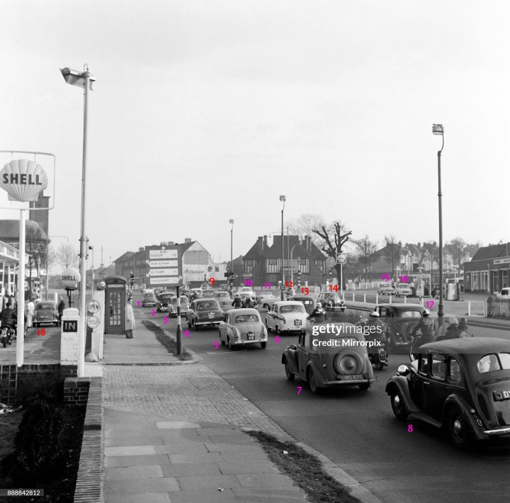 Pistonheads - This is a black and white photo capturing a bustling street scene in the United Kingdom. The image features multiple cars, each with their own unique design, navigating the busy streets. The vehicles are positioned in various ways, some appearing to be moving along the road while others seem to be parked or stationary. 

In addition to the cars, there's a gas station visible on the left side of the image, and a telephone booth located towards the right end. The presence of these structures suggests that this is an urban area with various amenities. 

The sky above is filled with clouds, adding to the overall vintage feel of the photo. Despite being in black and white, the image effectively conveys the hustle and bustle typical of a busy UK city street from the past. The perspective of the photo gives us a bird's eye view of the scene below, allowing us to see how tightly packed the cars are, hinting at heavy traffic or congestion.