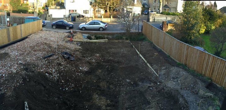 Our build thread, renovation and extension - Page 100 - Homes, Gardens and DIY - PistonHeads - The image depicts an excavation site with a large waist-high hole visible from the street level. Surrounding the hole is a wooden slat fence, which appears divided into sections. In the background, there are parked cars and residential buildings. The overall setting suggests that this could be a front or backyard of a house undergoing construction or renovation. There are no people visible in the image.