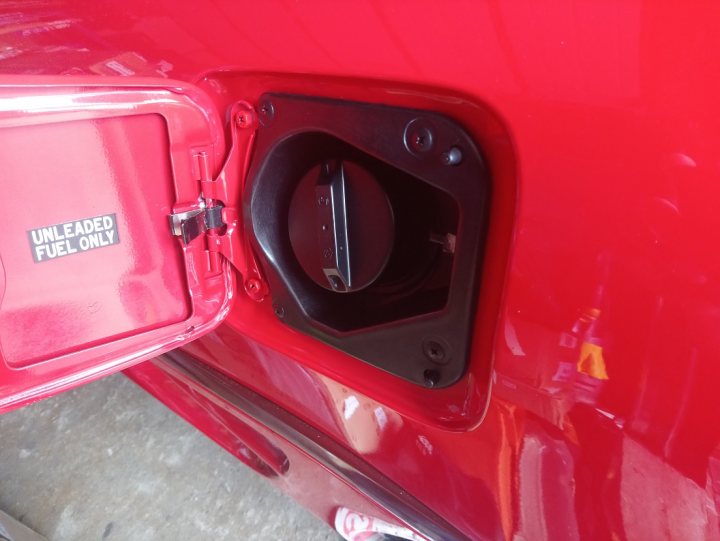 Low Mileage Toyota MR2 MK2. - Page 2 - Readers' Cars - PistonHeads - The image shows a close-up view of the fuel cap on a red vehicle. The cap is partially open, revealing a black handle which suggests that the gas tank is accessible. There is a white label with a barcode and text on it attached to the cap. The background is blurred but appears to be an indoor setting, possibly a garage or workshop.