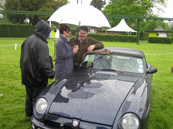 film cars - Page 1 - Classic Cars and Yesterday's Heroes - PistonHeads - The image presents a scene under a white tent where a small group of people are gathered around a dark blue car. One of them is leaning on the car's hood, while others are standing behind it. The handbag held by one of the individuals suggests they might be dressed for the day. The car displayed a Ferrari emblem, indicating it's a luxury vehicle. The people's clothing seems to be casual, which contrasts with the upscale nature of the car.