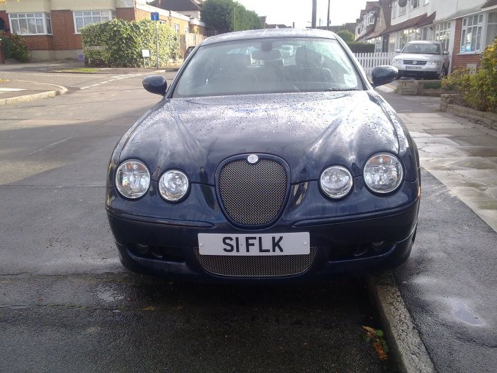 Twin Pistonheads Mustang Turbo - The image shows a dark blue Audi S4 on a wet street. The vehicle is parked parallel to the curb, its nose pointing towards the left of the frame, and displays a "SILFK" license plate on the front. In the background, other cars and buildings are visible under an overcast sky. The Audi has distinct lion badge emblems on the grille and fenders, indicating its series and make. There are lights turned on in neighboring houses, suggesting it might be evening or night time.