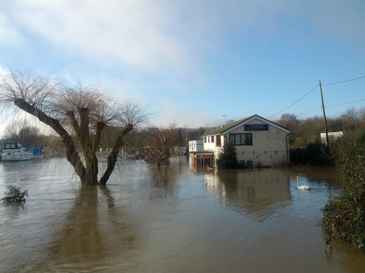 Maidstone town centre flooded - Page 1 - Kent & Essex - PistonHeads - The image showcases a scene of a flood. The main focus is a small house that seems to be affected by the rising water. The front part of the house, including the door, is submerged in the water. The sky above the house is cloudy and overcast, adding to the gloomy scene. The water appears to have washed away some vegetation in the foreground, creating a clearer view of the house. The surrounding trees seem to be wilted, possibly killed by the flood.