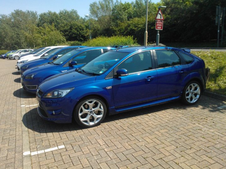 Who's got what in the South West - Page 1 - South West - PistonHeads - The image displays a row of diverse vehicles parked side by side. At the front and prominently displayed is a bright blue car, which occupies a significant portion of the image. The vehicle is parked over white lines on a textured, brick-style pavement. The background consists of a serene wooded area with trees and a glimpse of a grassy field or park. Small wildlife, likely a bird due to the triangular shape, can be spotted in the top left corner of the image. On the right side, there is a road with a triangular sign and a traffic light, indicating that the parking lot may be near a street or intersection. This scene suggests an outdoor setting, possibly a car show or public parking lot, given the orderly arrangement of the vehicles.