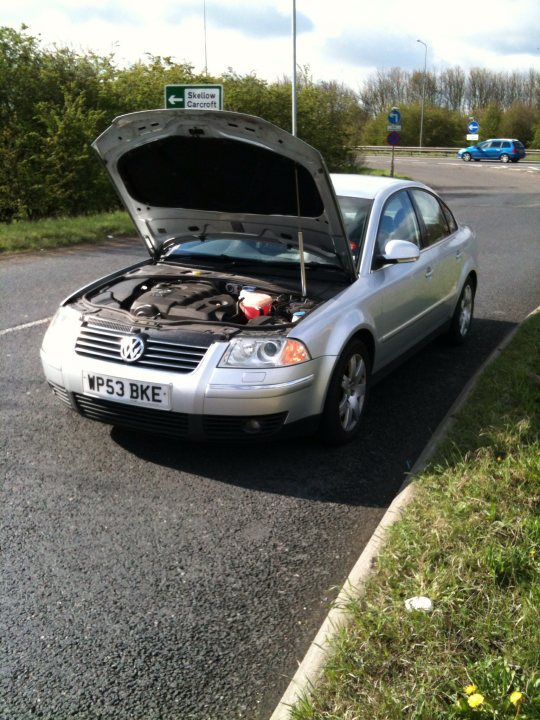 Garage blown my engine - where do I stand? - Page 3 - General Gassing - PistonHeads - The image shows a silver car parked on the side of the road within a lot. The car's hood is propped open, revealing the engine beneath. There's another car, a blue station wagon, driving in the opposite direction in the background. The scene appears to be set during the day, though the lighting suggests it might be overcast. The lot appears to be a parking area, and there are signs in the background that suggest it might be a public or commercial area. The car's condition, with the hood open and the engine exposed, might indicate that it's undergoing maintenance or that it has broken down and requires assistance.