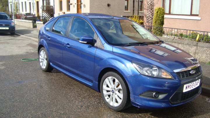 Aint Wait Corsa Currys Pistonheads - The image shows a parked blue car during a rainy day, as indicated by the reflection on the wet street. The car appears to be a compact, possibly a hatchback, and has two visible rear doors that may fold flat for added storage. On the windshield, there's a hint of resistance applied by the glass, suggesting it's recently rained or the weather is still clearing. To the left of the vehicle, a yellow hose extends onto the street, and the surrounding area includes other parked cars and a house with a visible window.