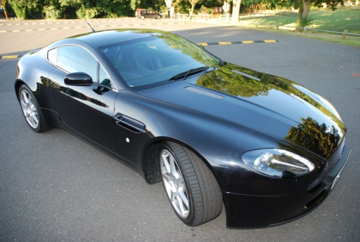 Pistonheads Promised - The image shows a sleek, black two-door sports car parked on an asphalt lot. The car's design is modern, with a prominent front grille and curved edges around the headlights and side mirrors. The car's side features two prominent front doors with a shorter rear door. The car is positioned at an angle to the camera, with the front of the car facing towards the right of the image. The reflection of the car on the ground suggests that the scene is bright and sunny, enhancing the glossy appearance of the vehicle.