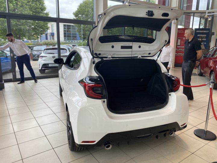 A white car parked next to a parking meter - Pistonheads - The image shows an indoor car showroom. At the center of the room is a white car with its trunk open, revealing luggage compartments filled with various items. A man in a suit stands behind the car, presumably explaining or demonstrating the vehicle. In the background, other vehicles and personnel can be seen, suggesting a sales environment. The room has a sleek design with white walls and floors, which creates an open and airy atmosphere.