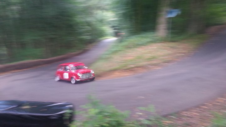 Pistonheads - The image shows a red classic car in motion on a winding country road. The car is moving towards the right side of the frame, with its body slightly leaning into the turn. The background features lush green trees, indicative of a rural setting, and there appears to be a patch of grass or dirt near the edge of the pavement. There's also a black vehicle parked on the left, partially obscured by the car in motion. The image is captured from a perspective that gives the viewer an elevated look at the scene.