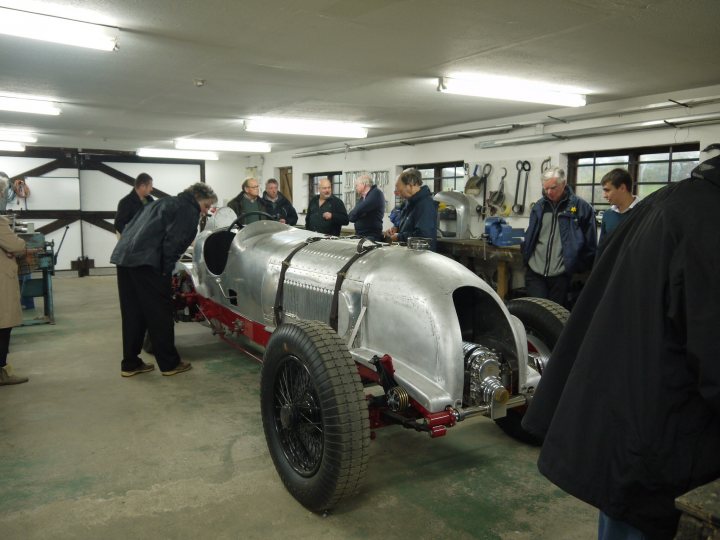 So what have you done with your Aston today? - Page 109 - Aston Martin - PistonHeads - This image captures a group of people gathered in a large room with a high ceiling. The focus is on a vintage car, which is silver with some red accents, and which occupies a significant portion of the room. The people, most of whom are men, are standing around the car, inspecting it, and engaging in conversation. The car itself is striking, giving off an air of antiquity and elegance that contrasts with the industrial feel of the warehouse-like setting. The lighting is subdued, casting soft light over the whole scene and highlighting the metallic sheen of the car.