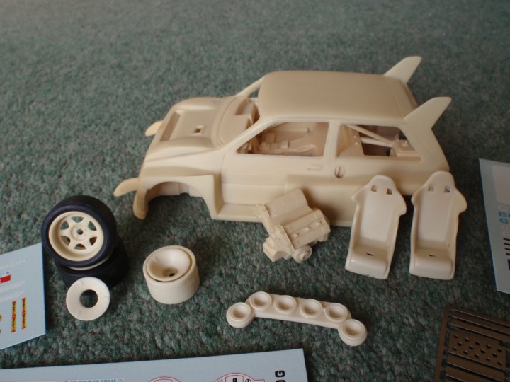 Metro Scale Pistonheads - This image captures an assembly in progress, with the remaining parts of a dollhouse car scattered on a carpeted floor. The car, which is the centerpiece of the scene, has already been partially assembled. The accompanying instruction manual stands open nearby, potentially guiding the assembly process.