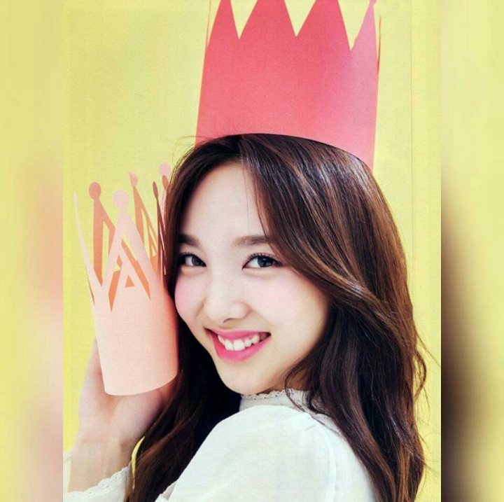 A woman holding a pair of scissors in her hands - A young woman smiling brightly at the camera, wearing a coral-colored paper tiara. Behind her, in her arms, is another similar tiara. She has long brown hair and is dressed in a white top. The background is yellow, which complements the coral color of the tiara. The image has a cheerful and joyful vibe.