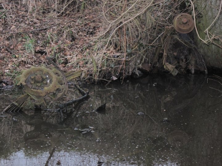 Any ideas what it is? - Page 1 - Classic Cars and Yesterday's Heroes - PistonHeads - The image shows a body of water reflecting a dead tree and moss-covered rusty metal objects. Among the objects, there appears to be a metal EKG machine lying in the water. The scene is mossy and the water is murky with reflections of the surrounding vegetation and debris. The forest interior suggests a rural, undergrowth setting.