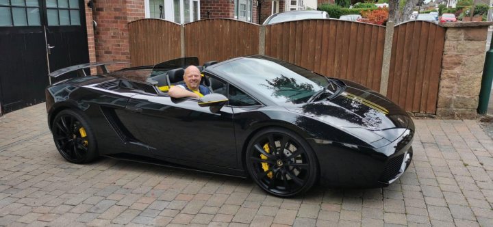 Bought myself a Bull - Page 1 - Readers' Cars - PistonHeads UK - The image shows a man sitting inside a black sports car with a convertible top. The vehicle appears to be parked in front of a brick wall and a gate, suggesting a private property or an exclusive area. The man is not wearing a mask, indicating that the photo was taken during a time when health precautions were less strict. He seems to be posing for the photo, as he is facing directly towards the camera. There's no visible text in the image.