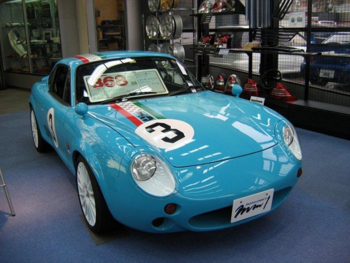 Shooting Metal Pistonheads Conversion Brake Roof - This image features a striking blue sports car with a race number 36 displayed on its hood. The car is predominantly blue, highlighted with a white stripe on the side and a contrasting white roof. It is parked in an indoor setting, possibly a showroom or a museum, as indicated by the various models of cars in the background. The lighting in the room illuminates the car, capturing its sleek design and vibrant color. The contrast between the blue car and the neutral background enhances the car's presence in the scene.