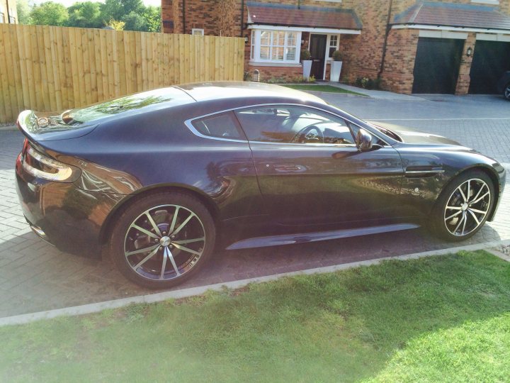 So what have you done with your Aston today? - Page 107 - Aston Martin - PistonHeads - The image showcases a black Aston Martin with a sleek design, parked at the end of a driveway. The car features a glossy finish, with shiny rims adding to its luxurious appeal. The driveway itself is of concrete, leading up to a residence in the background. The scenery suggests a quiet, suburban setting with a well-maintained lawn and trees in the distance.