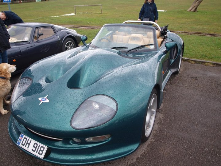 Marcos Display - Clevedon 14/04/2013  - Page 1 - South West - PistonHeads - The image shows two vintage sports cars parked side by side on what appears to be a wet, paved surface. Each car has its own unique style and color, with one being blue and the other black. The cars' rounded shapes and prominent headlights suggest a design from the 1970s or 1980s. There is a hint of a person standing behind the cars, but the focus of the image is on the automobiles. Interestingly, one of the cars displays a license plate with "918", which could be a reference to the Japanese anime and manga series "Neon Genesis Evangelion", though this is speculative without further context.