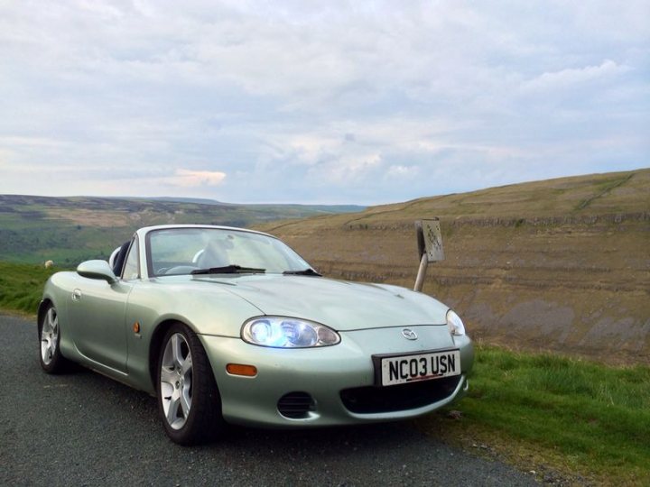 my mx5 - Page 1 - Mazda MX5/Eunos/Miata - PistonHeads - In the image, a blue sports car with a California license plate is the focal point, parked by the side of a country road. The road leads through a picturesque landscape, with a grassy hill and a clear sky above. On the other side of the road, there is an open field meeting a hill, providing a serene background. The car's sleek design and vibrant color stand out against the natural scenery, indicating a sense of adventure or leisurely travel.