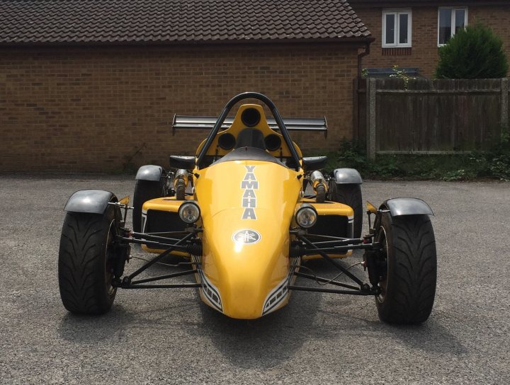 Roadrunner Racing SR1 - Page 1 - Readers' Cars - PistonHeads - The image showcases a sleek, primary yellow race car situated in a driveway, most likely belonging to a house with two visible windows. The car's design is characterized by its aerodynamic shape, complete with visible aero parts and a rear spoiler, suggesting it's designed for speed and maneuverability. It features two seats, one visible in the image, and displays a black front bumper which contrasts with its yellow body. The gravel driveway hints at a possibly rural or semi-rural setting, away from paved roads.