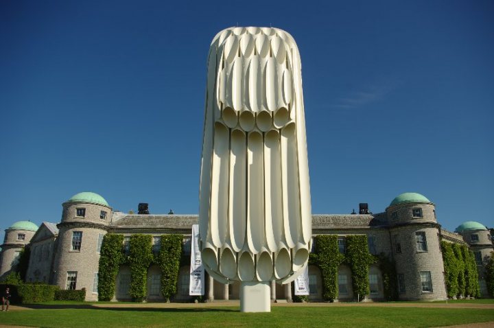 Anyone been to the Moving Motorshow before? - Page 1 - Goodwood Events - PistonHeads - The image features a large, white sculpture with a curved section at the bottom. It stands prominently in the foreground, with lush green foliage in the background. In the distance, there's a stone building adorned with several dome-like structures on its roof. The sky above is clear and blue.