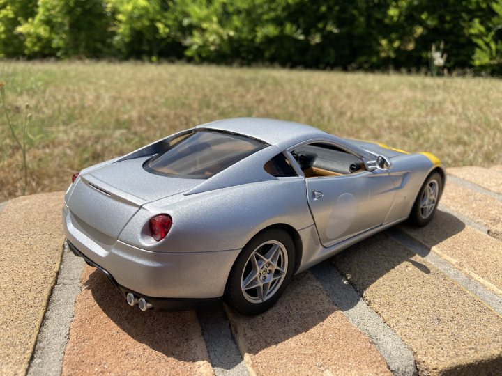 A blue car parked in a parking lot - The image shows a small, silver toy car with a convertible top parked on a brick surface. The vehicle is detailed and realistic in appearance, suggesting it may be a model or an actual toy designed to resemble a real sports car. In the background, there's a lush green lawn under a clear sky, indicating that the photo was taken outdoors during daylight hours. A person can be seen in the distance, providing a sense of scale and context to the scene. The style of the image is a straightforward photograph with no filters or artistic manipulations evident.