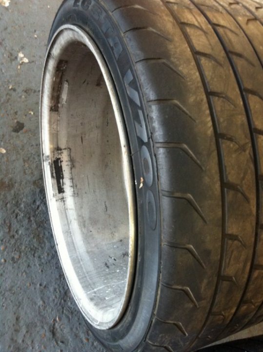 A blue and yellow fire hydrant on a sidewalk - Pistonheads - The image shows a close-up view of a tire from a vehicle, with a focus on the center tread where the words "Goodyear" can be seen. The tire appears to have been used, as there are signs of wear and dust. The rim of the wheel, which is white in color, reveals its damaged condition with visible dents and scratches, indicative of impact or forceful data. The background is an indistinct, grey, rough concrete surface, suggesting the tire might be in storage or on display.