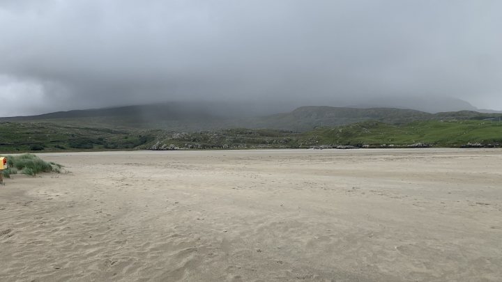 Southern Ireland 7 day driving tour. - Page 1 - Holidays & Travel - PistonHeads UK - The image shows a serene beach scene. There is an overcast sky, suggesting it might be a cloudy day. The sand of the beach is visible, along with some grass and possibly rocks or pebbles, indicating that this beach may have areas where the tide goes out, leaving exposed land. A small figure in a red jacket can be seen walking towards the water's edge from the foreground, providing a sense of scale to the landscape. In the distance, there are mountains with clouds hanging over them, adding depth to the scene. The overall atmosphere is one of tranquility and natural beauty.