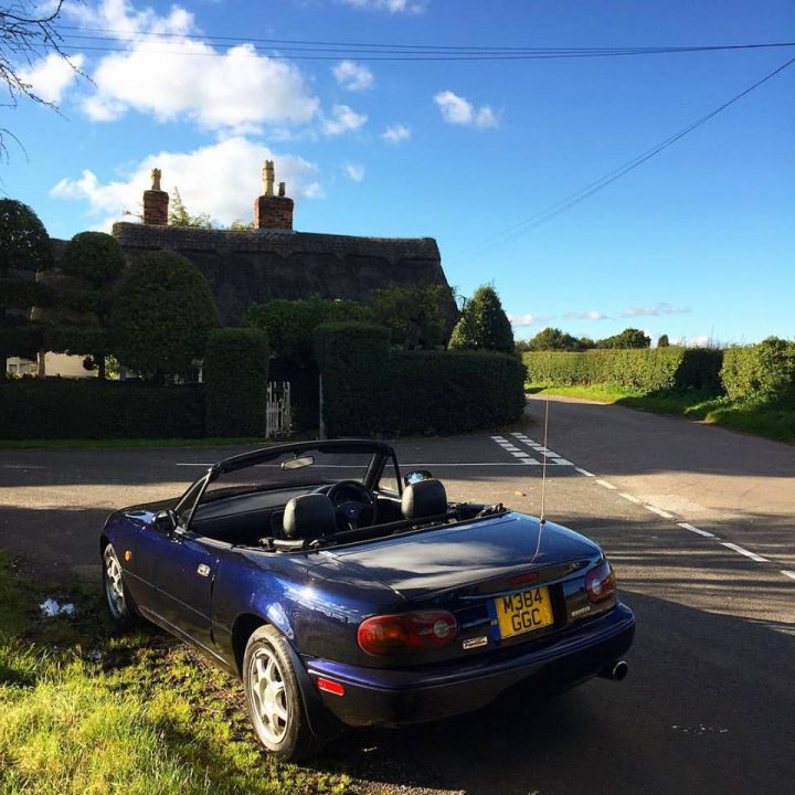 Uh oh I'm going to look at my (potential) 3rd MX5 tonight... - Page 1 - Mazda MX5/Eunos/Miata - PistonHeads - The image shows an outdoor scene with a road that divides the picture. On the right side, alongside the road, is a large, well-maintained mansion with a steeply pitched, dark roof and prominent chimneys. The sky above is blue with scattered clouds, suggesting a clear day. In the foreground on the left side of the image, a blue sports car is parked. The car has a convertible top down, allowing glimpses of its interior, and it is positioned at an angle that suggests it might be facing forward, as indicated by the rear licence plate which reads "M33 GGC." The road itself is marked with white lines and a dashed line indicates a divided path. The overall impression is of a serene roadside location, likely at a crossroads, adjacent to a substantial residential property.