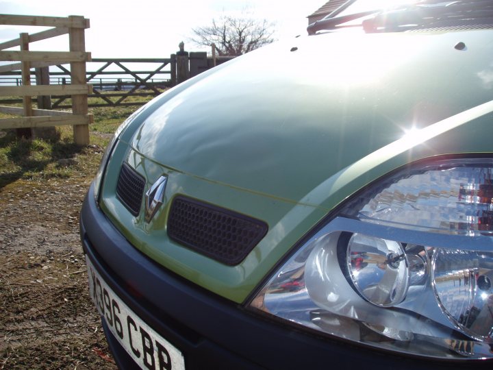 my first car  - Page 1 - Readers' Cars - PistonHeads - The image depicts the front of a small green car parked on a gravel road near a wooden fence. The vehicle is positioned at half an angle to the camera, highlighting the front headlight, bumper, and grille that features a chrome emblem. The sky is clear, suggesting a sunny day. The setting appears to be in an open rural area.