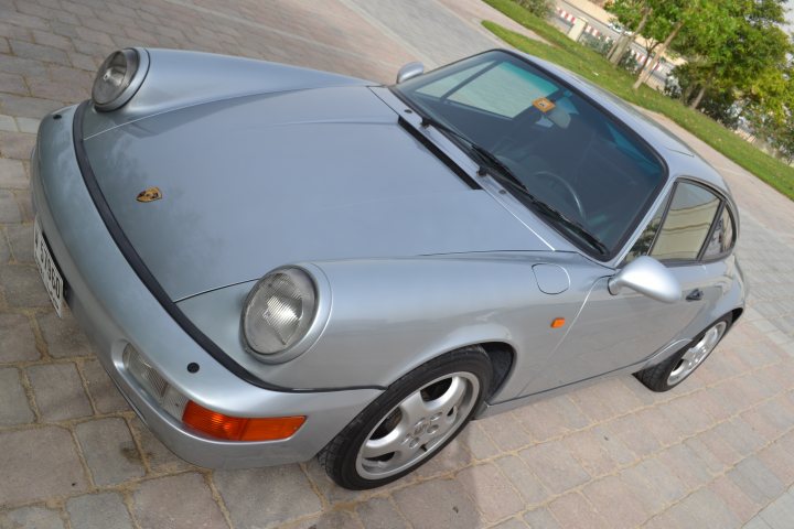 RE: The easiest job in the world? - Page 3 - General Gassing - PistonHeads - The image captures a single, classic silver convertible car parked on a brick parking lot. The vehicle's hood is raised slightly, revealing its engine, and a matching black softtop is folded down, giving a sense of its convertible nature. The car has the Porsche logo and badge, indicating it's a Porsche model. The surrounding environment includes a green lawn, trees, and a cloudy sky, providing a peaceful and natural backdrop to the parked luxury vehicle.