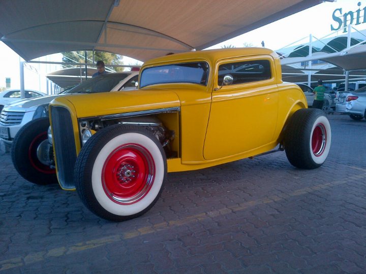 Middle East spotted thread - Page 64 - Middle East - PistonHeads - The image depicts a vintage yellow car parked in a lot. The antique vehicle has red and white accents on the hubcaps, and it appears to be a classic model with a distinct design. The car is positioned under a structure with a "Spirit of America" sign, suggesting it might be parked in a dedicated area for classic vehicles at an American-themed venue. In the background, there are other cars and benches, indicating that this is a public location. The setting seems to be a sunny day, as indicated by the bright lighting and shadow cast by the car.