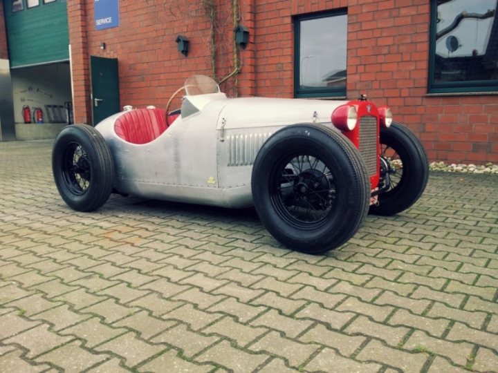 Talk to me about Austin 7 Specials - Page 1 - Classic Cars and Yesterday's Heroes - PistonHeads - The image presents a vintage red and white racing car parked on a brick driveway in front of a red brick wall. The car appears to be in good condition, with its large wheels and prominent grille. There is a green sign high up on the brick wall, but its contents are not clearly legible in the picture. In the background, there is an open door that offers a glimpse into a room, partially visible through the windows on either side of the door.