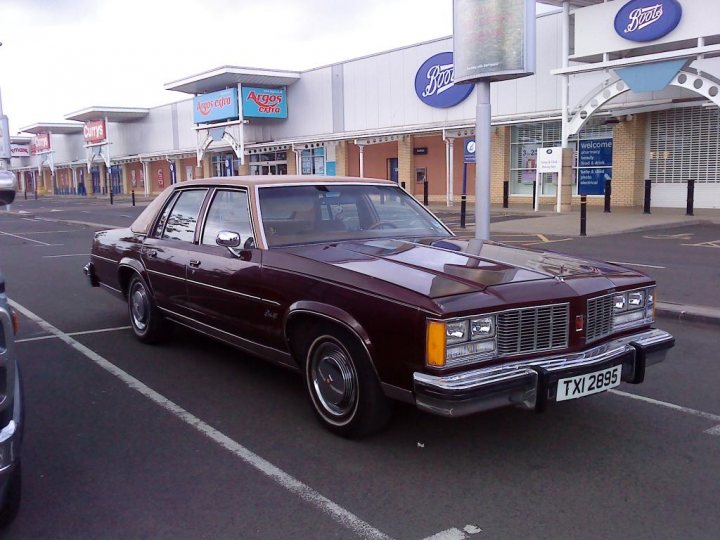 Cruisemeet Pistonheads Edinburgh - The image shows a dark red, four-door luxury sedan parked in a parking lot with marked spaces. The car is of a vintage design, given away by its curved roofline and overall shape. It's pulled up to the corner of a space and is parked adjacent to a narrow sidewalk. The background features a shopping area with various stores and signage, including a Pizza Hut sign. The sky is overcast, suggesting an overcast day.