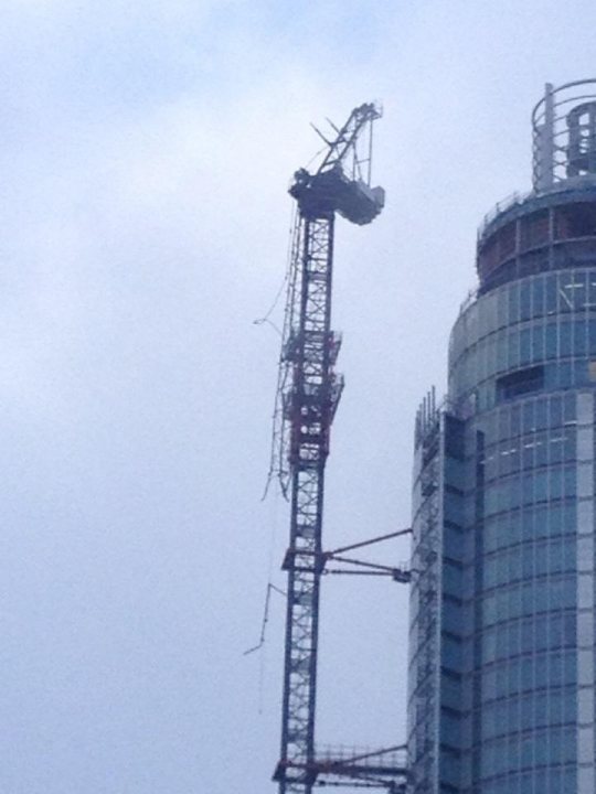 A large building with a clock on it - Pistonheads - The image shows a large, multilevel tower with a prominent, unfinished section that appears to be a construction site. Atop the tower, there is a large, circular silver object resembling a water tank. Below this circular structure, there are several levels of metal beams and scaffolding that indicate active construction or renovation. The sky is overcast with a slight blue peek, suggesting an overcast day.