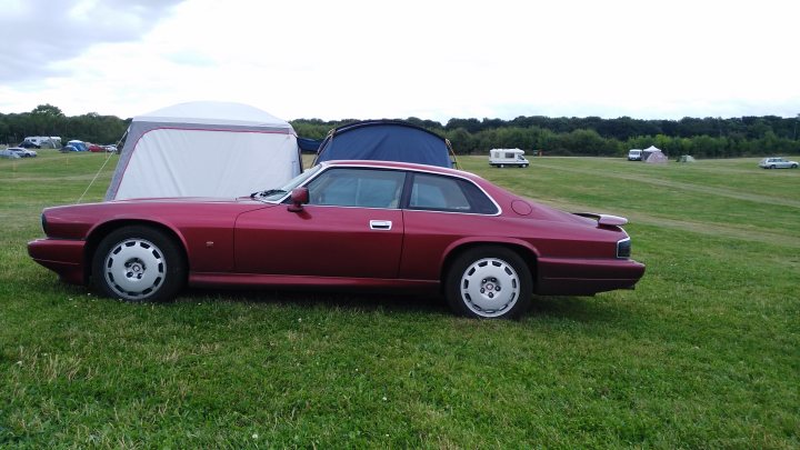 1992 Jaguar XJR-S - Page 5 - Readers' Cars - PistonHeads - The image features a striking red, two-door classic sports car parked on a grassy field. The car, equipped with a hardtop and fiberglass bug shield, is positioned with the front facing towards the left side of the frame. Behind the car, a red dome tent is set up on the same grassy plot, suggesting a camping setup. The field is surrounded by trees, providing a serene and natural backdrop for the vehicle and tent. In the distance, other vehicles can be seen in the far background, but the focus remains on the sports car and its accompanying set-up.