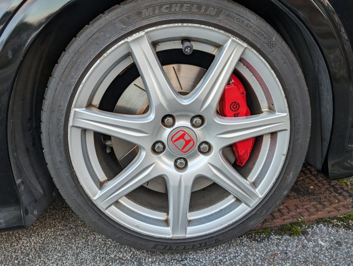 Civic Type R FN2 Track Car  - Page 10 - Readers' Cars - PistonHeads UK - The image is a close-up photograph of a silver-colored wheel on a car. It features the vehicle's front right tire with the wheel well visible, showcasing the design and hubcap of the alloy. The rim is multi-spoke, and there are red accents around the center cap. In the background, another part of the car is visible, suggesting that this image is taken from an angle close to the ground. There are no texts present in the image.