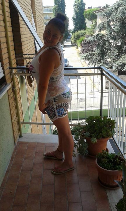 Vakantie - In the image, a woman is standing on a balcony. She's wearing a tank top and shorts, and appears to be looking at the camera with a slight smile. The balcony has a view of a street below, indicating that the building is likely an apartment or condo complex. The sky is clear, suggesting it might be late afternoon or early evening. There are potted plants on the balcony, adding a touch of greenery to the scene. A handbag can also be seen in the image.