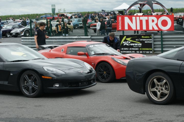 Things they dont tell you when you SELL an Elise - Page 1 - Elise/Exige/Europa/340R - PistonHeads - The image captures an event taking place at a venue with a racetrack. There are several luxury and sports cars parked on the tarmac, surrounded by people who appear to be enjoying the spectacle. One prominent sign in the scene reads "NITRO" which suggests the name of the event or a level of performance associated with the vehicles. There is another sign that mentions "KUPWER," and other people in the background, adding to the lively atmosphere of the event.