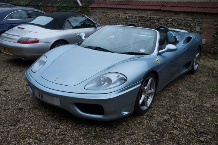 360 thread - Page 1 - Ferrari V8 - PistonHeads - The image presents a tranquil outdoor scene featuring three sports cars parked on gravel. The central focus of the image is a **mid-engine sports car**, painted in a metallic **blue color**, parked on top of the gravel. This particular car, a ** convertible**, is a **Ferrari**. The convertible top is down, exposing the car's interior and engine. In the background, two additional sports cars are visible, parked on the left side, one with a **dark blue color** and the other in **silver**. The gravelly ground and the sky make up the backdrop of the scene.