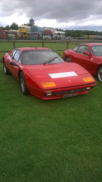 Classic Ferrari at Croft today. - Page 1 - Ferrari Classics - PistonHeads - The image features two vintage sports cars parked on green grass. The car in the foreground is a bright red Ferrari with the license plate "M50 BVY". The second car, parked behind the Ferrari, is of a similar red color but is an older model. Both cars have distinctive orange stripes on the front. In the background, there appears to be light traffic and commercial buildings, suggesting that the parking area might be in an urban or semi-urban setting. The overall scene conveys a nostalgic atmosphere.