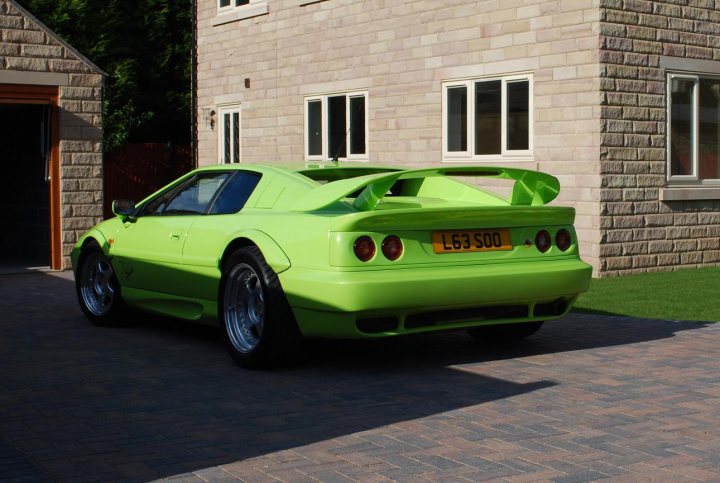 Pictures of my Sport 300 at last! - Page 1 - Esprit - PistonHeads - The image displays a bright green sports car parked in front of a two-story brick building. The car has a black soft top convertible roof and a large rear wing, indicative of a modified sports car. There are three windows visible on the building. The vehicle is parked on a brick driveway, suggesting a residential setting. The sunlight casts a strong shadow on the brick pavement.