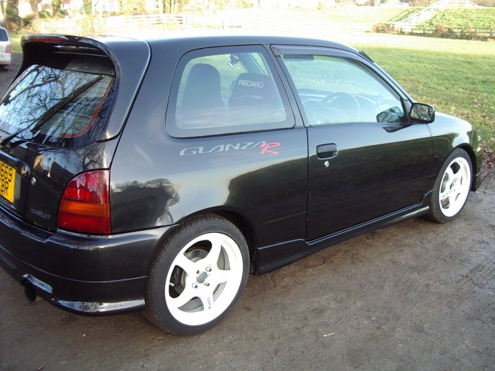 Toyota Starlet/Glanza - Page 1 - Jap Chat - PistonHeads - The image captures a scene of an aging black Mozart parked on a gravel surface. The once synchronized red and white decal on the driver's side door is slightly faded, bearing signs of age and weathering. The car is positioned to the left of the frame, with a grassy area visible behind it. A lone headlight on the right side of the car stands like a sentinel against the backdrop of undulating terrain.