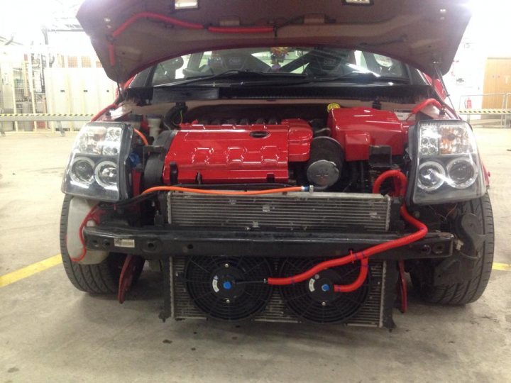 A motorcycle is parked in a parking lot - Pistonheads - The image shows a scene in a garage or workshop, focused on a red car that has the hood open. Its impressive engine, which includes various components visible through the open hood cover, is disconnected from its engine block. Multiple wires and a loose belt are tangled around the car's body, suggesting a complex repair or maintenance work in progress. The vehicle's design features a prominent front grille and curved headlights. The garage has a yellow line on the floor, which is typically used to mark a boundary for safety purposes. The overall setting implies a professional environment dedicated to automotive work.