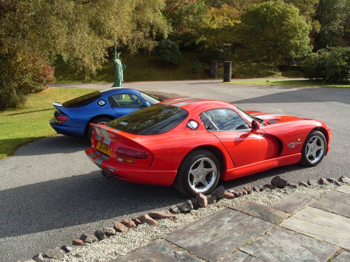 scottish gts meet - Page 1 - Vipers - PistonHeads - The image captures a tranquil scene of two sports cars parked on a gravel driveway. One car, a striking red sports car, is camouflaged with white stripes. A bit further away, a blue sports car is parked, complete with a large American flag decal. The vehicles are situated in front of a lush, grass-covered hillside, adding a touch of nature to the scene. The concrete area where the cars are parked is dotted with a few rocks. The overall atmosphere is quiet and serene.