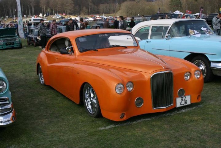 'Different' Jaguars. - Page 11 - Classic Cars and Yesterday's Heroes - PistonHeads - The image depicts a gathering in a grassy field at a car show. There are numerous vintage cars on display, with one particularly striking car, featuring a bright orange paint scheme with a classic black roof. The classic design elements, including round headlights, a grille with horizontal bars, and fender-mounted mirrors, retroact precisely to mid-20th-century American styling. Surrounding the show cars, there are people milling about, some standing near the vehicles while others are scattered in the distance. The field itself appears well-maintained, with trees dotting the background, and the overall atmosphere is that of a community event celebrating the classic and the vintage.