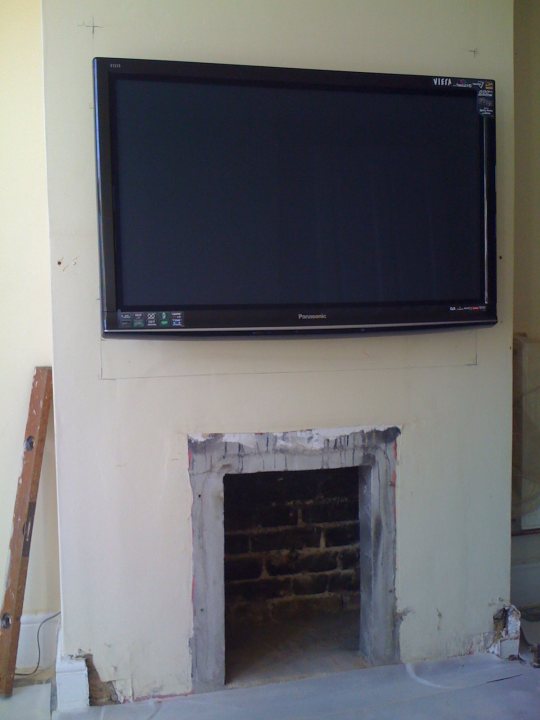 Tvhome Annoy Cinema Haters Pistonheads Lounge - A television mounted on a wall above a fireplace. The television has a flat-screen design and is in an off state. The room appears to be undergoing some renovations or alterations, as suggested by the unfinished look of the wall and the presence of a ladder in the room. The fireplace has an old-fashioned brick design, and the surrounding area is bare and gray.