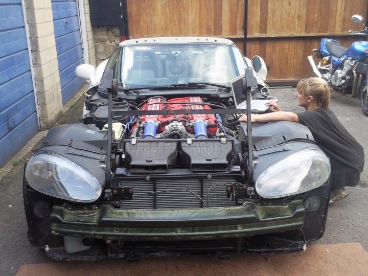 Rebuild Viper Pistonheads - The image shows a young person sitting underneath the hood of a car, surrounded by various mechanical parts, presumably engaged in some sort of maintenance or repair work. The car appears to have its engine compartment open, and the tender seems focused on the task at hand. There is another person visible in the background, possibly another mechanic or an observer. The setting appears to be an outdoor garage or a similar workspace, with a motorcycle parked to the side. The atmosphere suggests a practical, hands-on learning environment.