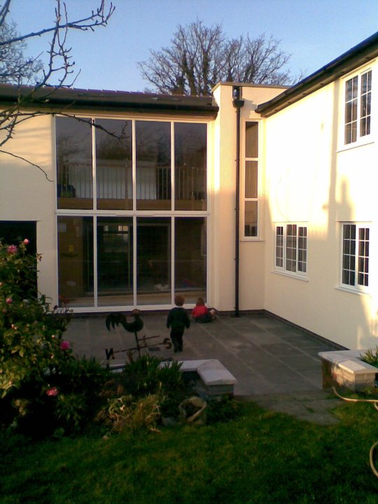 What does your house look like? - Page 5 - Homes, Gardens and DIY - PistonHeads - This image captures a charming scene of a family enjoying a sunny day outside their two-story house. The house features large windows that provide plenty of natural light. A tall, black downspout runs down the front of the residence, diverting water away from the building. The front yard is well-maintained with a beautifully manicured lawn. A young child can be seen happily sprawled out on the concrete patio area, soaking up the sunlight. The atmosphere appears peaceful and idyllic, typical of a perfect sunny day at home.